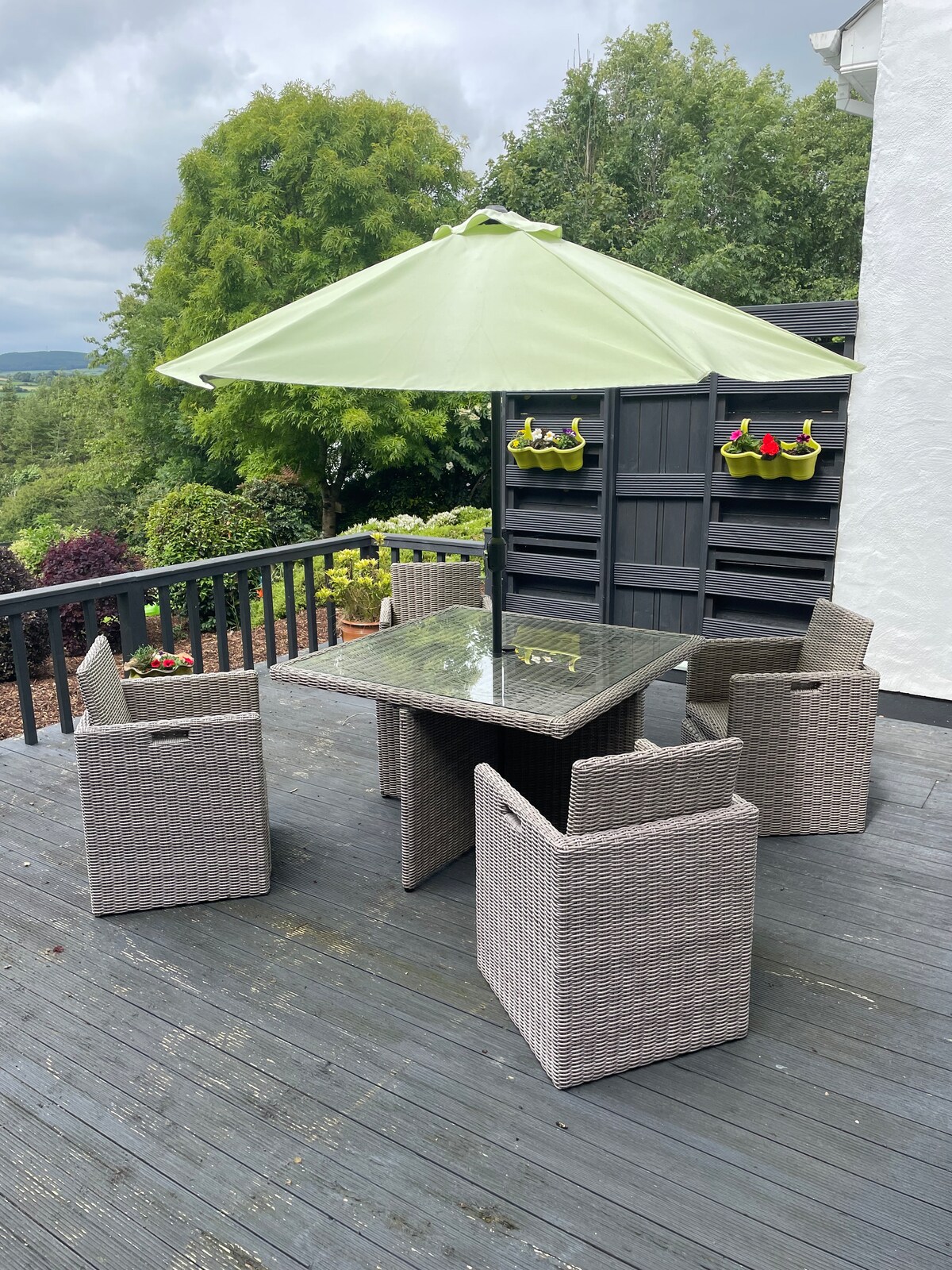 A private patio deck features a glass-top table surrounded by four wicker chairs, all situated beneath a light green umbrella. The space is framed by a dark wooden railing and is complemented by potted flowers on a nearby wall.