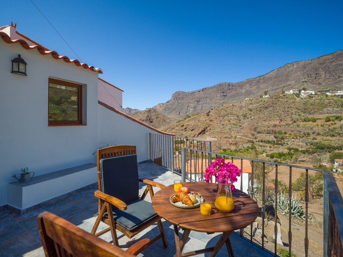A terrace features a round wooden table set with breakfast items, including pastries and drinks. Two chairs are positioned for comfort, with vibrant flowers providing a pop of color. Scenic views of the surrounding mountains create a tranquil backdrop.