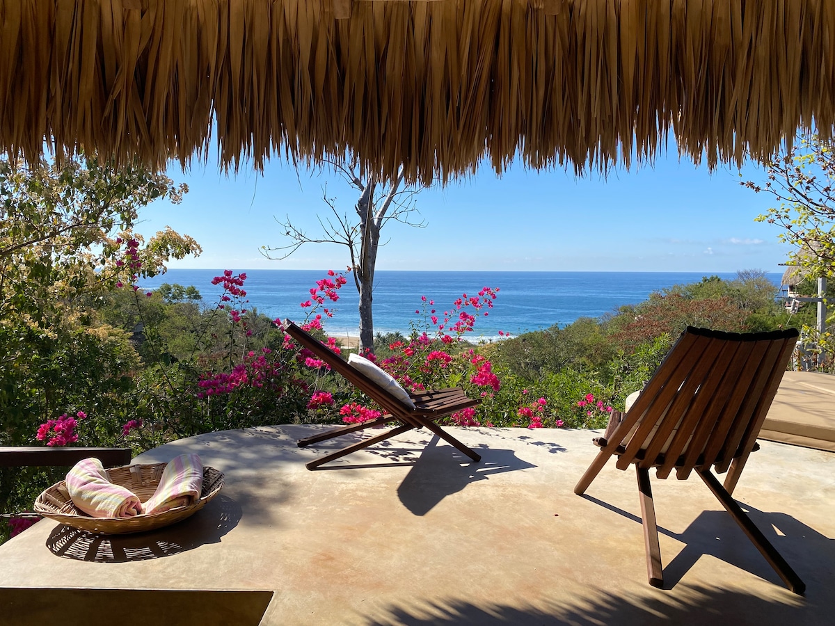 An outdoor seating area features two wooden chairs positioned on a terrace, overlooking a vibrant tropical landscape with blooming flowers. The Pacific Ocean is visible in the distance, framed by greenery and a thatched roof overhead.