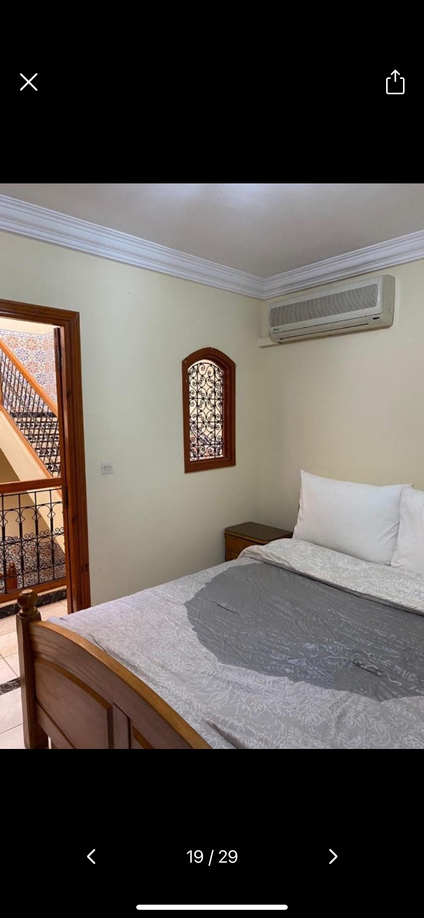 A well-lit bedroom features a spacious bed with white pillows and a light gray blanket. A wooden bedside table is positioned next to the bed. A window with decorative grill work adds architectural interest, while an air conditioning unit is mounted on the wall.