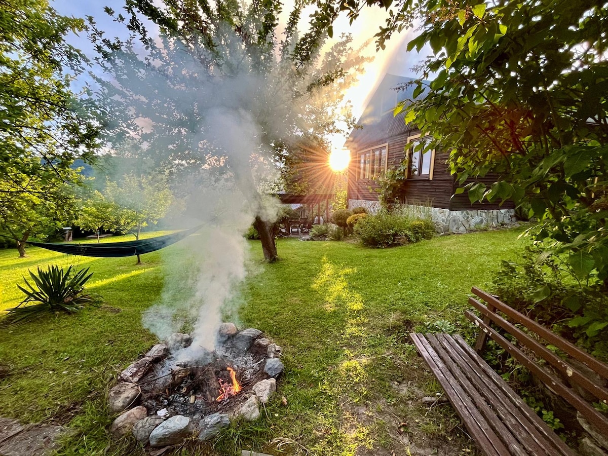 A cozy outdoor area features a small fire pit with smoke rising gently, surrounded by stones. Lush green grass and trees are visible, with sunlight filtering through the leaves, creating a serene atmosphere. A bench invites relaxation nearby.