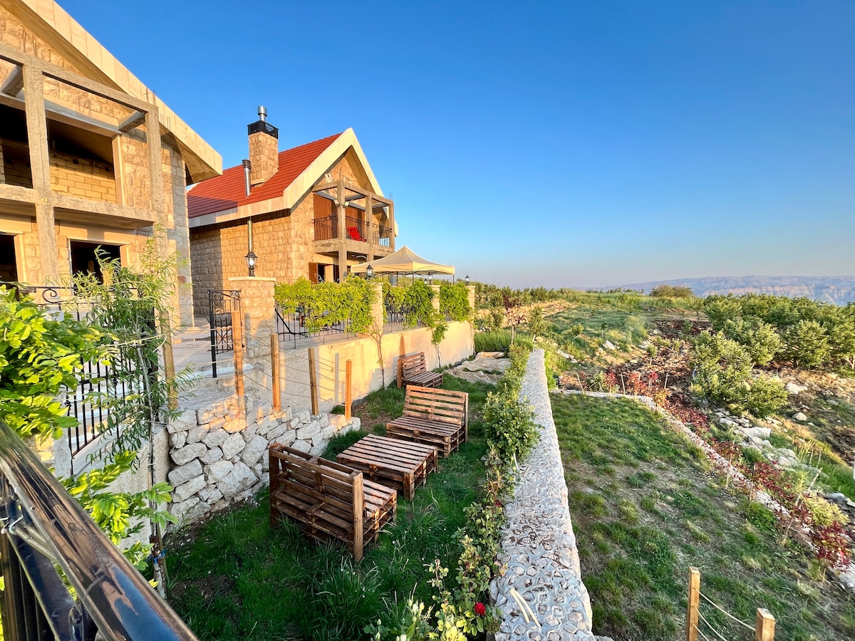 A stone chalet with a red roof is set against a clear blue sky, surrounded by lush greenery. In the foreground, wooden lounging benches are arranged on a stone patio, offering a tranquil outdoor space. The rolling hills in the distance create a serene backdrop.