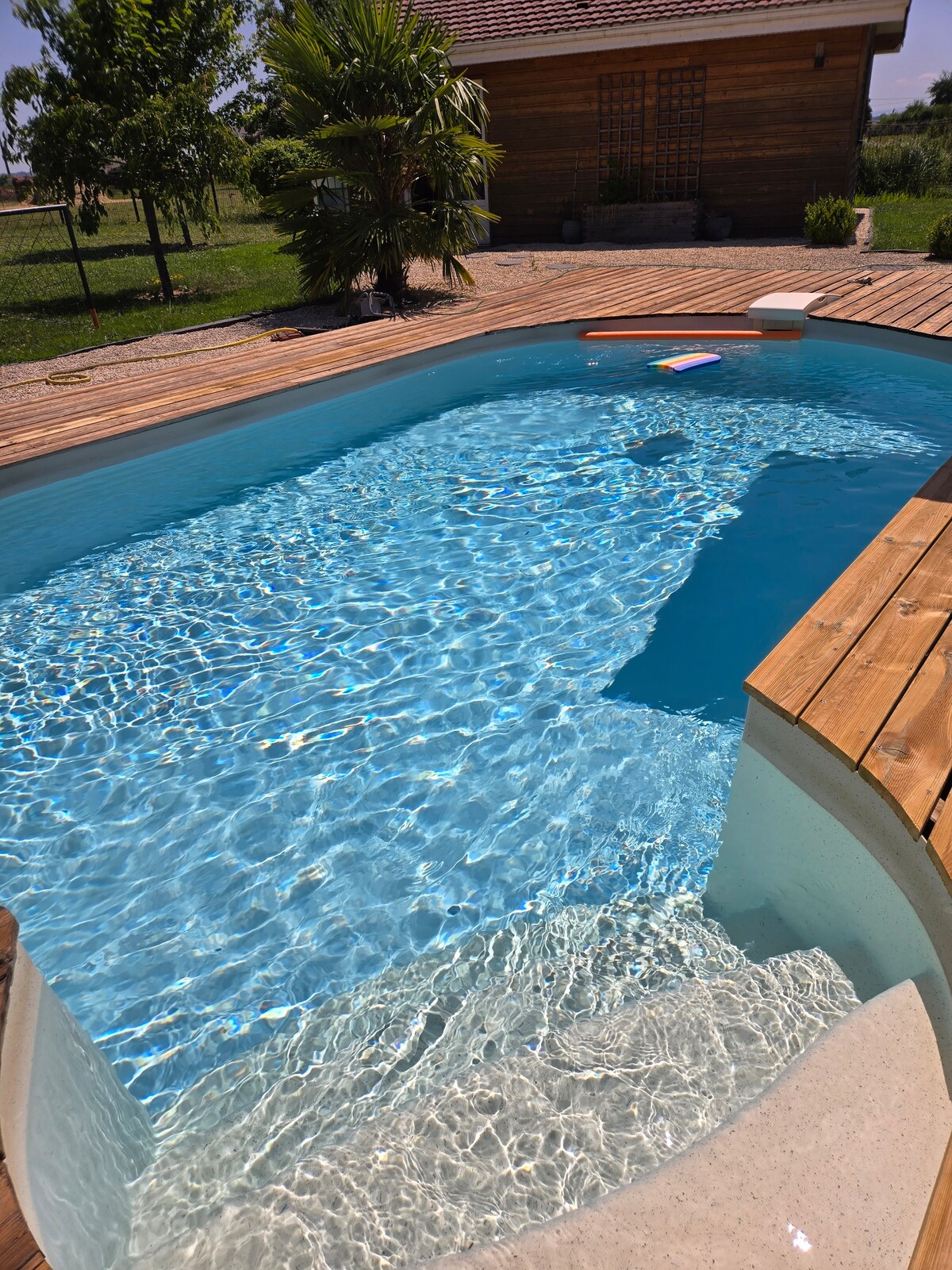 A well-maintained swimming pool is framed by wooden decking, reflecting sunlight on its clear water. The pool features a gentle slope leading to shallow water, and lush greenery is visible beyond the deck, enhancing the outdoor environment.