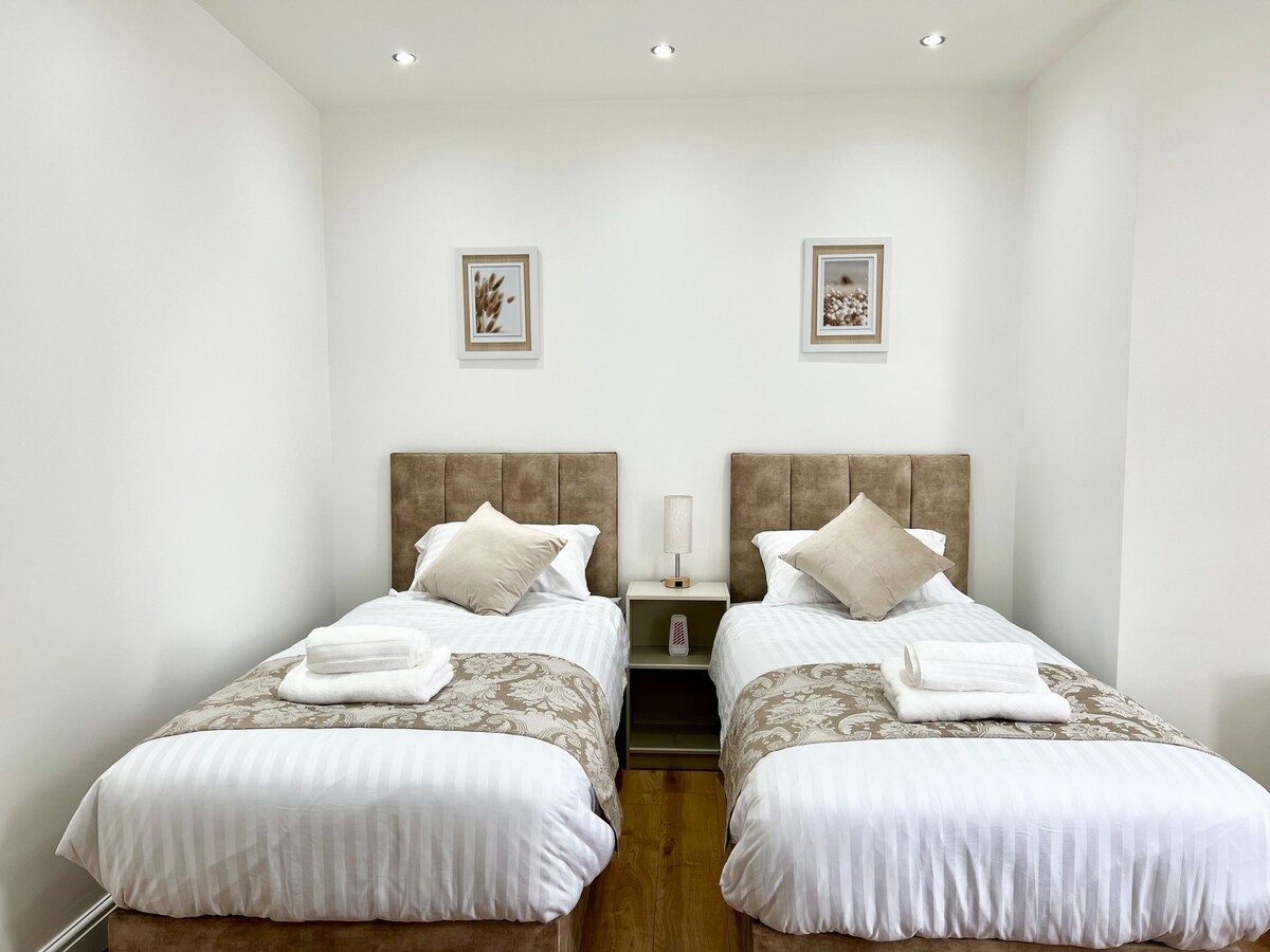 A brightly lit bedroom features two extra-long single beds with neutral bedding and decorative pillows. Bedside tables with lamps are positioned between the beds. Artwork adorns the walls, and natural light fills the space, creating a warm and comfortable atmosphere.