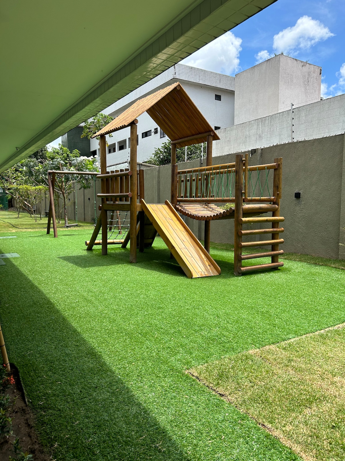 An outdoor play area features a wooden structure with a slide, rope bridge, and climbing elements, situated on a manicured grassy lawn. The space is surrounded by greenery and has a clear blue sky above.
