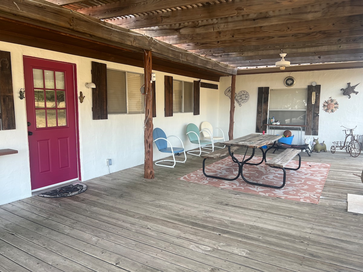 A covered patio area features a round table with bench seating and four chairs in light blue and white. A pink door provides a vibrant entrance, while wooden beams and a patterned rug add texture to the outdoor space.