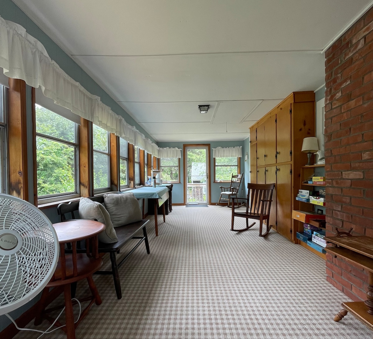 A bright sunroom features large windows allowing natural light to fill the space. Comfortable seating options include a rocking chair and a sofa. A fan is positioned nearby for added comfort. A wooden cabinet and bookshelves offer storage and reading materials.
