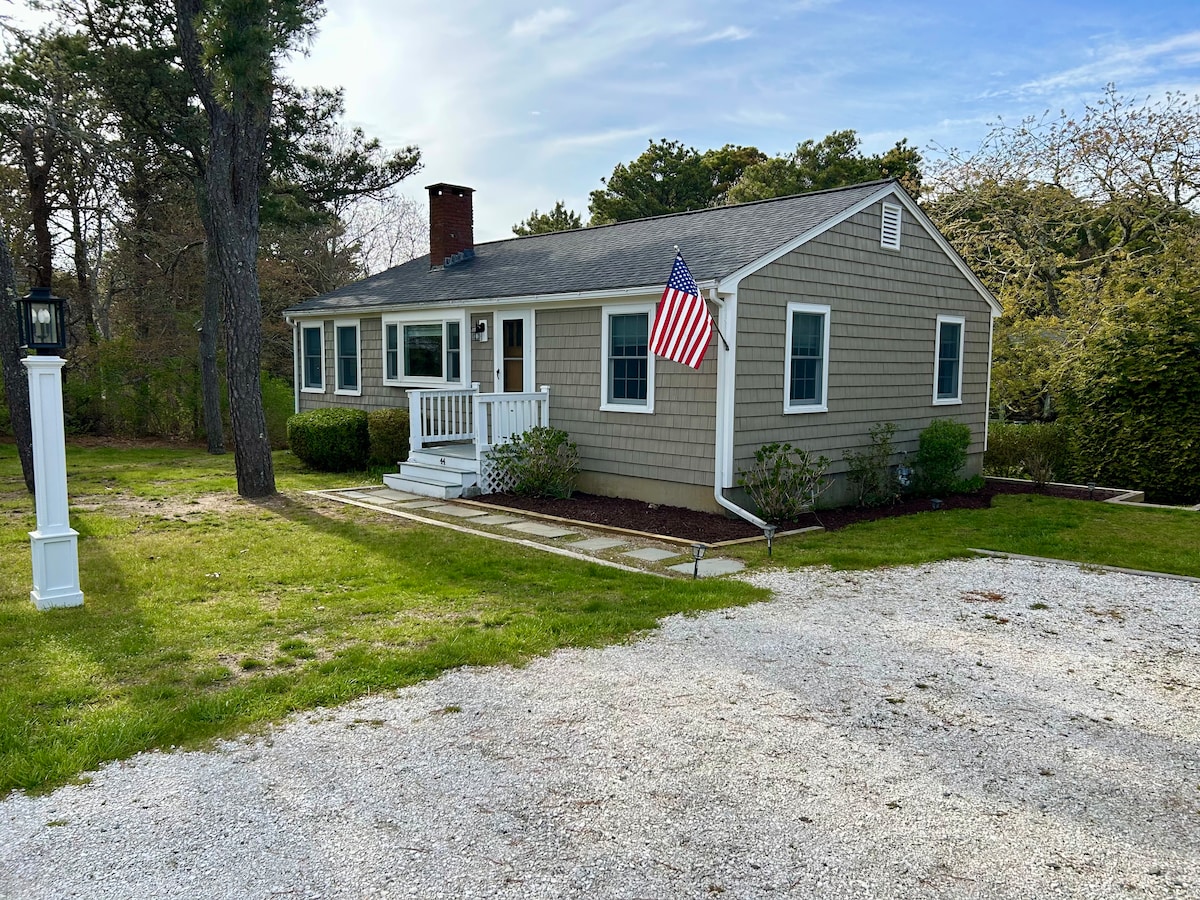 A charming cottage is situated on a gravel driveway, framed by greenery and featuring a flagpole adorned with the American flag. A front porch with steps leads to the entrance, complemented by well-kept landscaping and a peaceful atmosphere.