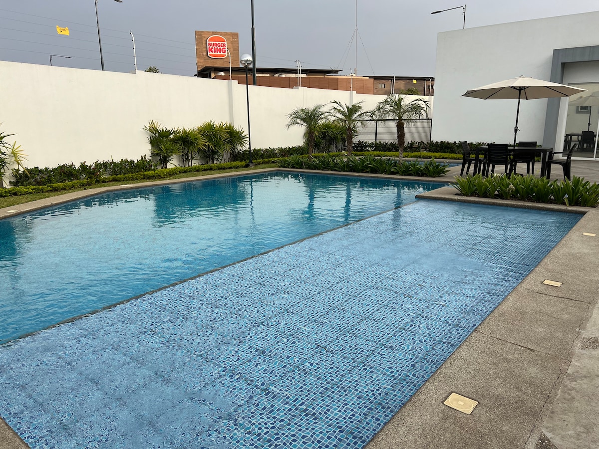 A clean and spacious pool area is presented, featuring two distinct swimming pools surrounded by lush green landscaping. A shaded seating area with a dining table and an umbrella is visible, providing a relaxing outdoor space.