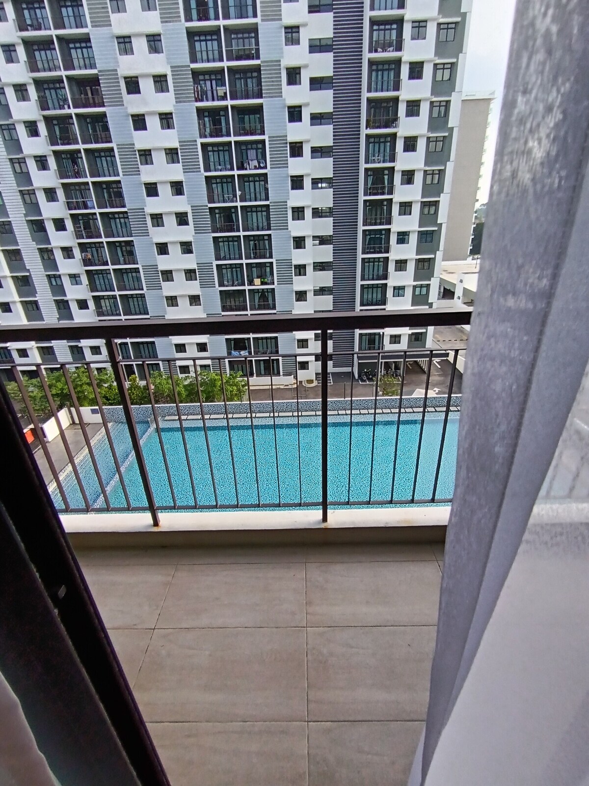 A balcony provides an exterior view of a turquoise pool surrounded by greenery, with an expansive apartment building visible in the background. The scene is framed by sheer curtains, enhancing the tranquil setting.
