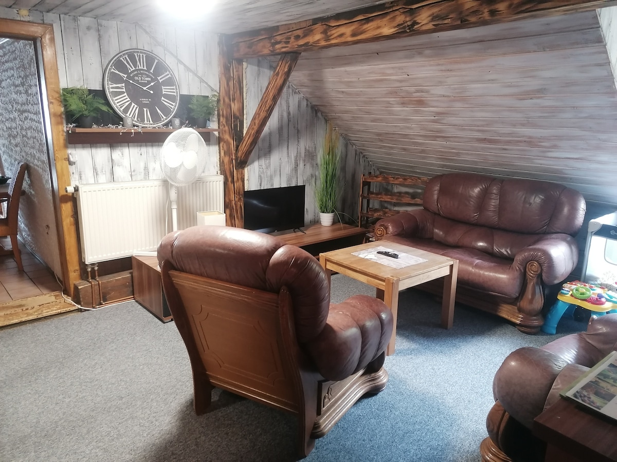 A cozy living area is presented with wooden beams and a sloped ceiling. Two brown leather sofas are positioned around a wooden coffee table. A flat-screen television is situated near the wall clock, and a small fan adds comfort to the space.