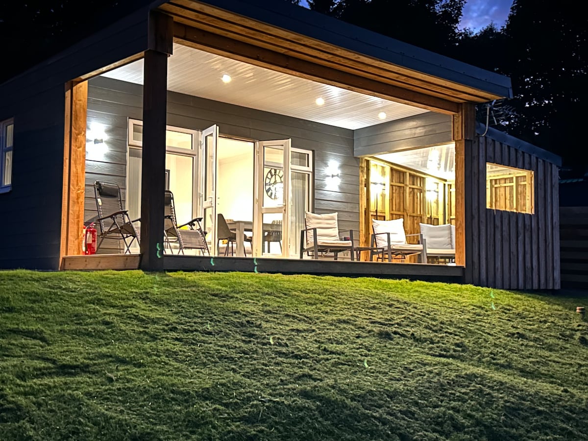 The holiday lodge features a spacious and modern design with large glass doors that open to the outdoor area. Comfortable seating arrangements are visible inside, complemented by warm lighting. The manicured lawn slopes gently towards the lodge, enhancing the open and welcoming atmosphere.