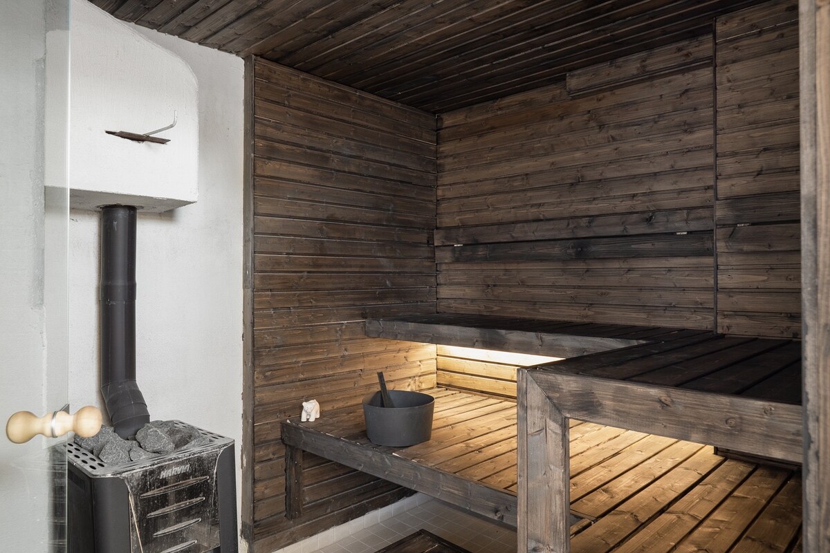 A sauna room is presented, featuring wooden benches and slatted walls, creating a natural, warm environment. A stove with stones sits against one wall, with a bucket placed on the bench, indicating a space ready for relaxation.