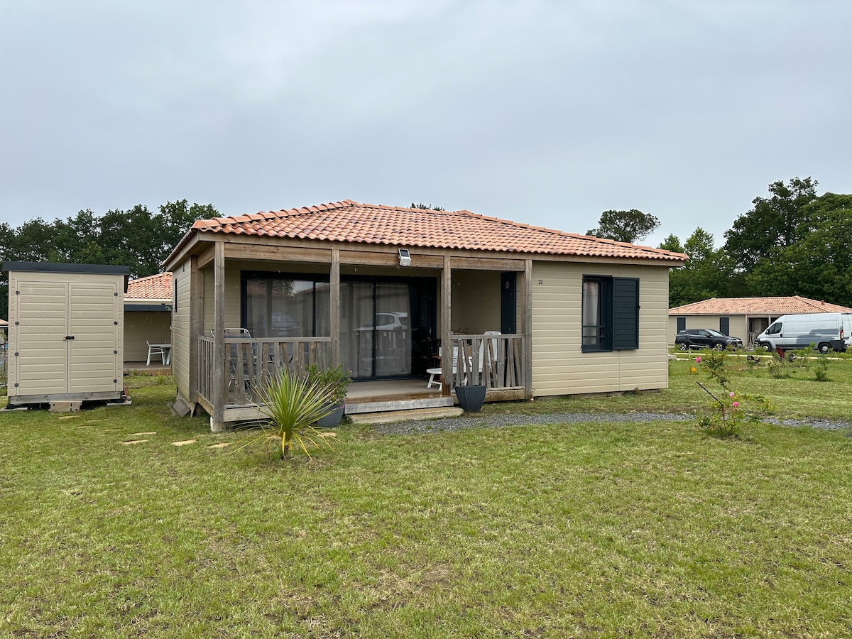 A single-story cabin with a tiled roof is surrounded by a grassy area. The porch features wooden railings, and large glass doors offer views into the interior. Natural light is visible through the windows, enhancing the inviting exterior.