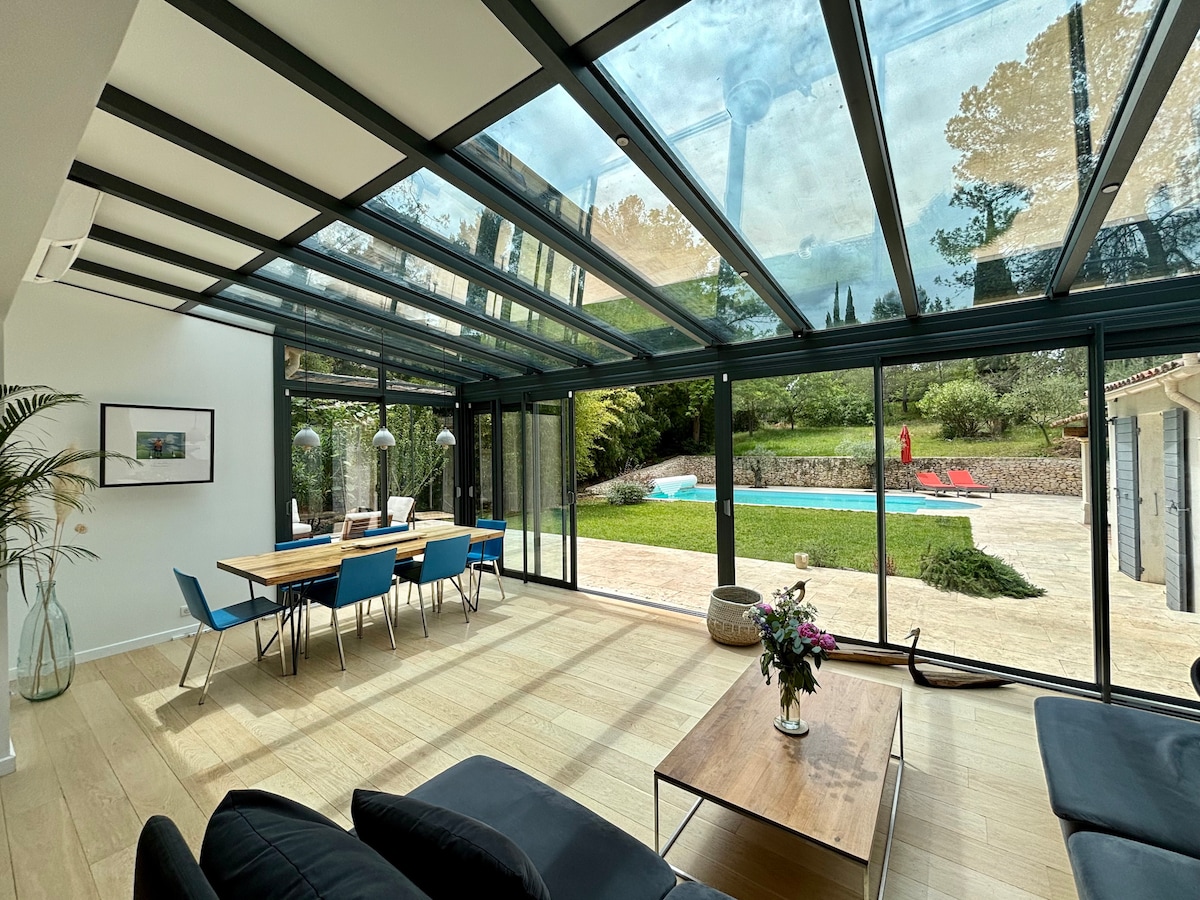 A spacious living area features floor-to-ceiling glass walls, providing a view of the lush outdoor garden and inviting swimming pool. A dining table with blue chairs accommodates several guests, while a coffee table rests in front of a comfortable seating area.