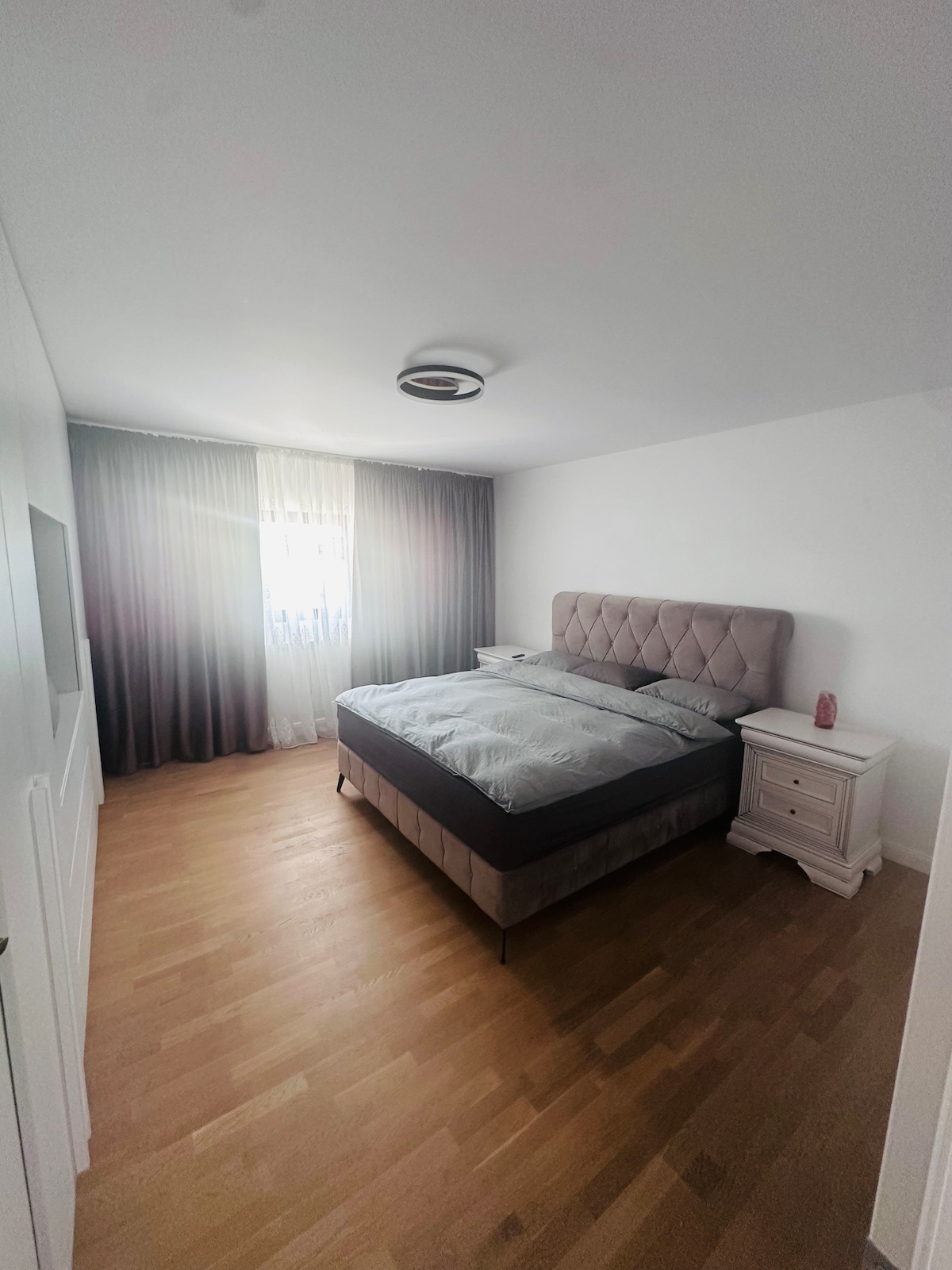 A well-lit bedroom features a spacious bed with a tufted headboard. Soft bedding is arranged neatly, and natural light filters through sheer curtains. A nightstand with a small decorative item stands next to the bed, while warm wooden flooring complements the room's serene ambiance.