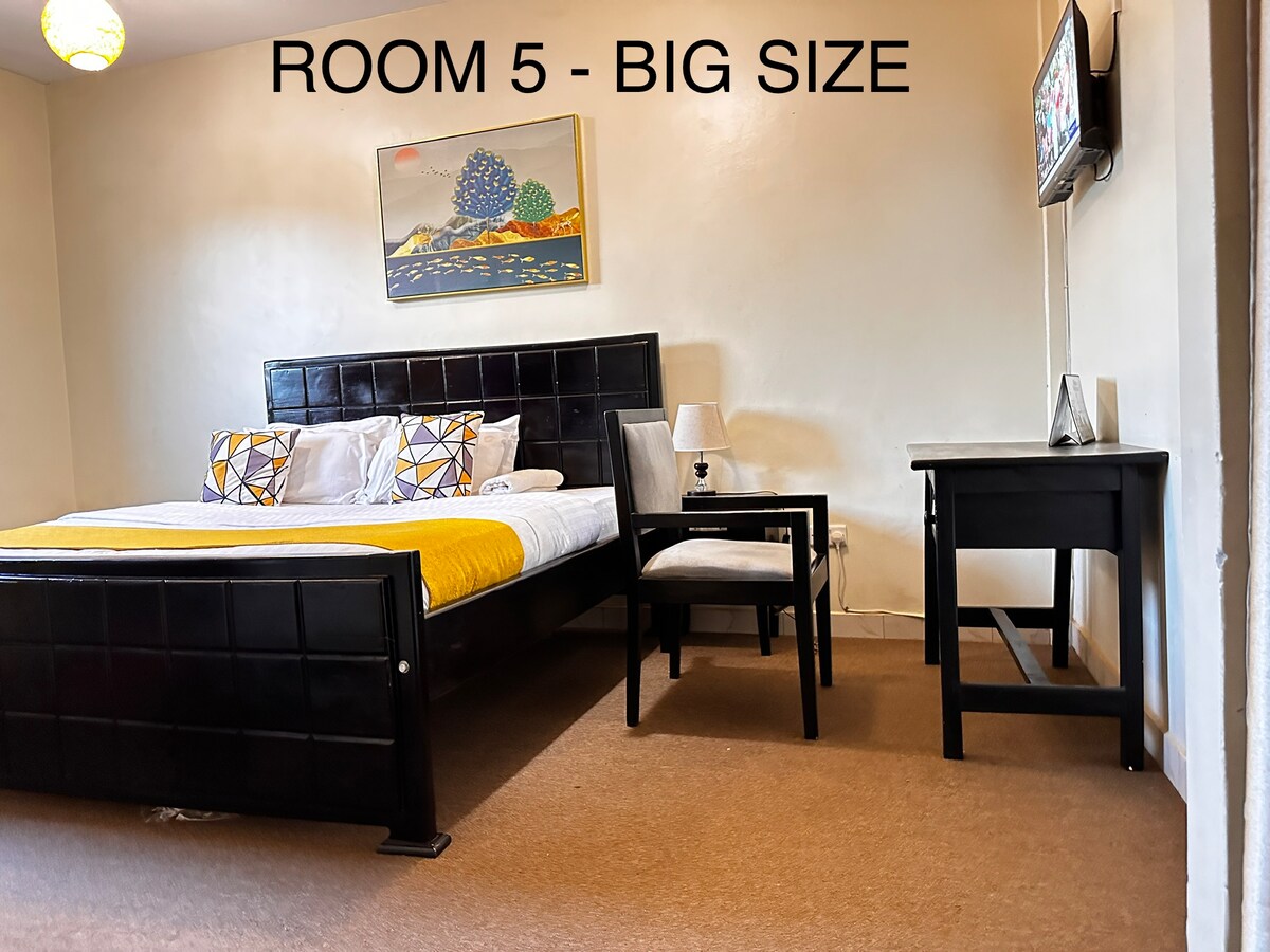 A spacious room features a large bed complemented by decorative pillows. A small desk and chair are positioned against the wall, while a flat-screen TV is mounted nearby. Natural light enters through a window, and a piece of wall art adds a touch of color.
