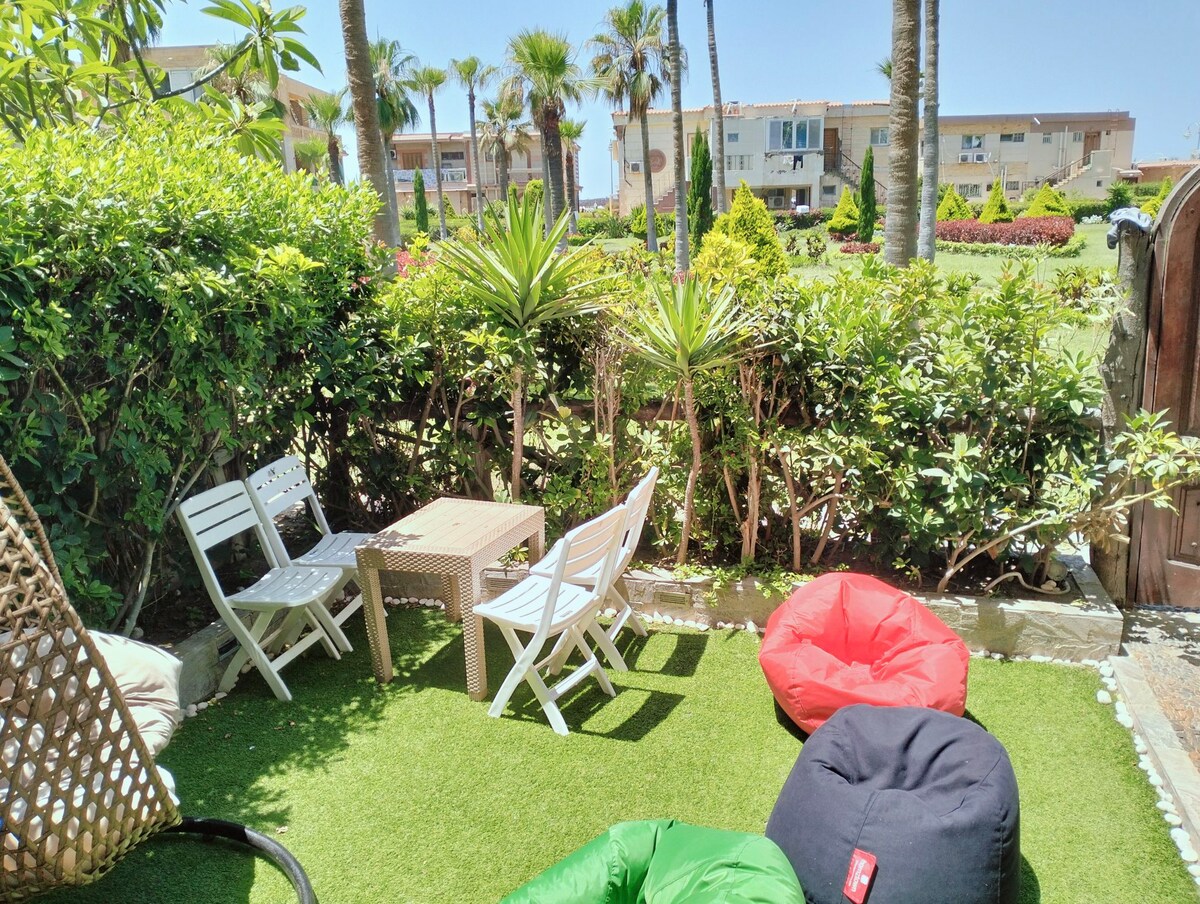 A private garden area is depicted, featuring several seating options including two white chairs and a wooden table. Colorful bean bags are arranged on the green grass, surrounded by lush greenery and palm trees, creating a relaxing outdoor space.