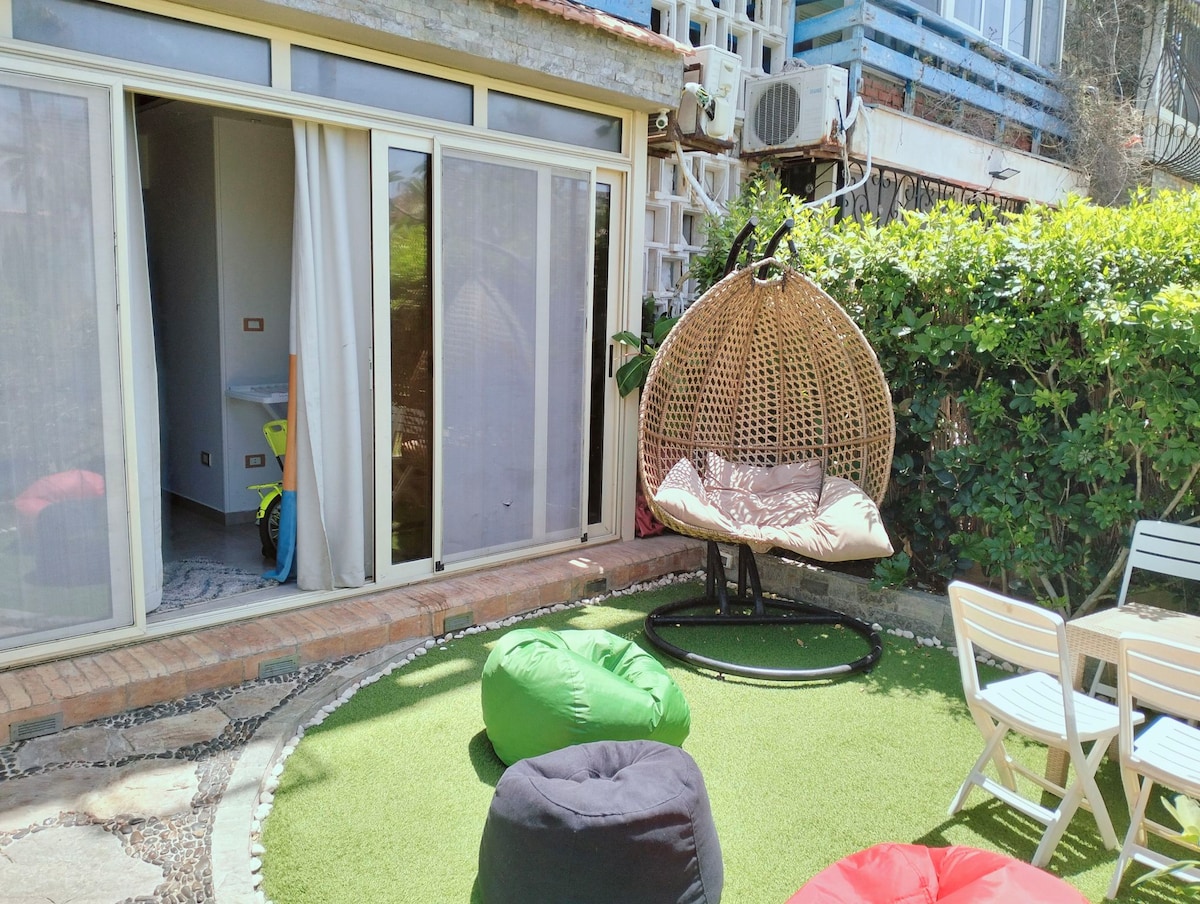 An outdoor area features a hanging chair and colorful bean bags on a manicured lawn. Close to the seating, large glass doors provide access to an indoor space, with greenery enhancing the privacy of the garden.
