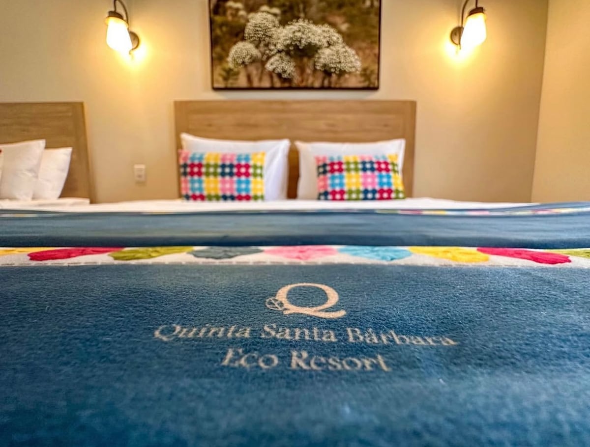 The image showcases a well-appointed sleeping area featuring a vibrant, colorful blanket laid over a spacious bed. Pillows with varied patterns rest atop the headboard. Soft light emanates from wall-mounted sconces, illuminating a decorative wall art piece in the background.