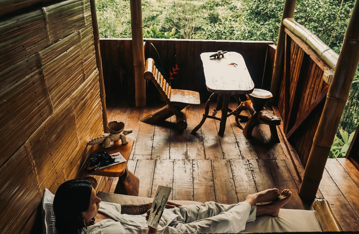 A cozy outdoor space features bamboo furnishings, including a table and two chairs, creating a perfect spot for relaxation. Lush greenery is visible through the open railing, enhancing the serene jungle ambiance. The wooden deck provides a warm and inviting setting for unwinding with a good book.