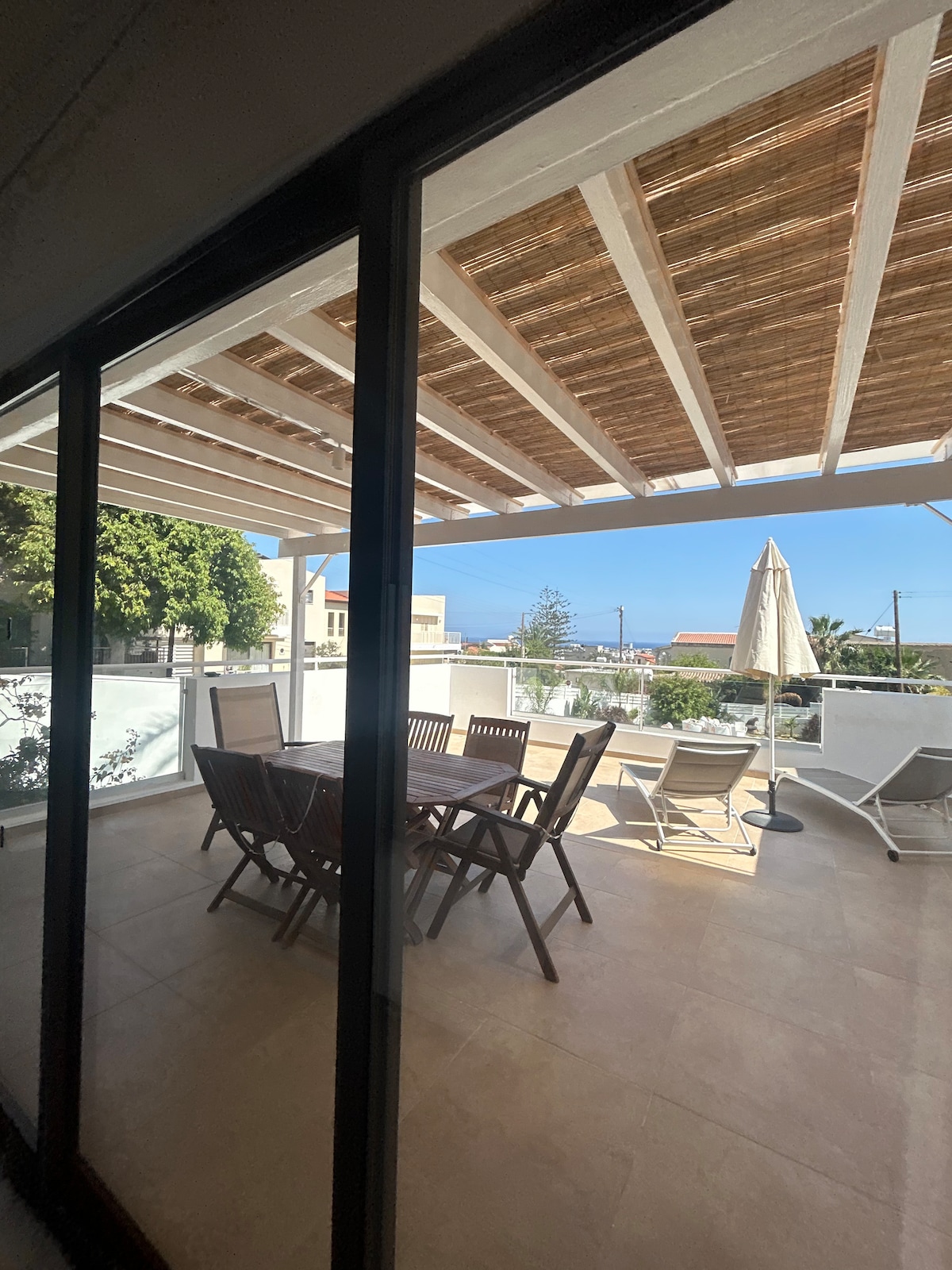 A spacious balcony is showcased, featuring a large dining table with wooden chairs and sun loungers beneath a bamboo-covered pergola. Views of greenery and the blue sky are visible through sliding glass doors, creating an inviting outdoor space for relaxation.
