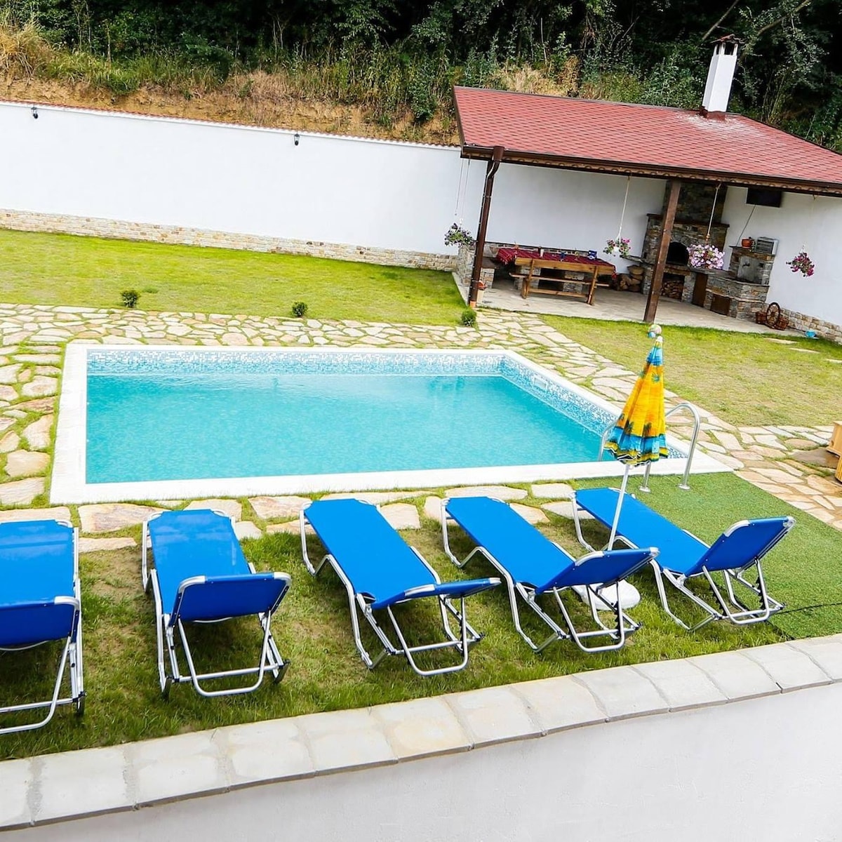 An outdoor area features a crystal-clear swimming pool surrounded by a stone patio. Multiple blue lounge chairs are set on the manicured lawn, with a colorful umbrella providing shade. In the background, a covered seating area is visible, designed for relaxation and social gatherings.