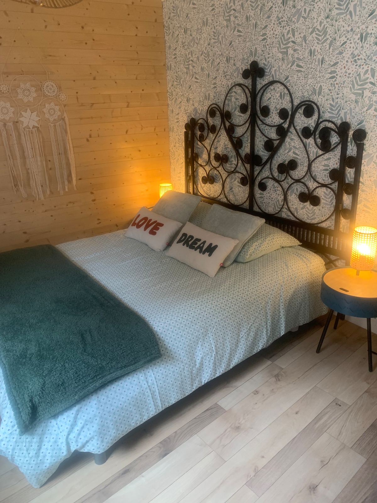 A double bed is centered against a wooden wall, adorned with a decorative headboard. Two patterned throw pillows with the words 'LOVE' and 'DREAM' are placed on the bed. Side tables with lamps are positioned on either side, softly illuminating the cozy sleeping area.