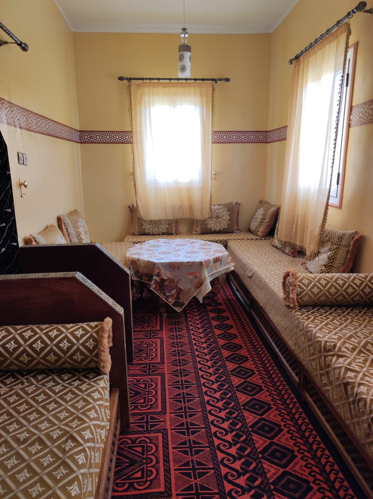 A cozy sitting area features ornately designed fabric-covered seating arranged around a central table. Light streams through a window with flowing curtains, creating a warm ambiance on the tiled floor, which is decorated with an intricate pattern.