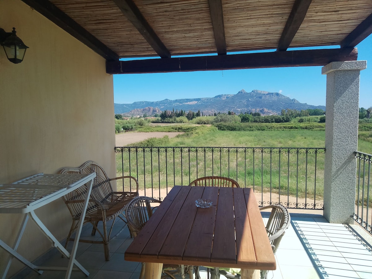A covered terrace features a wooden table surrounded by woven chairs, offering a panoramic view of green fields and distant mountains. A separate folding table is visible to the left. Soft sunlight illuminates the area, creating a serene outdoor space for dining.