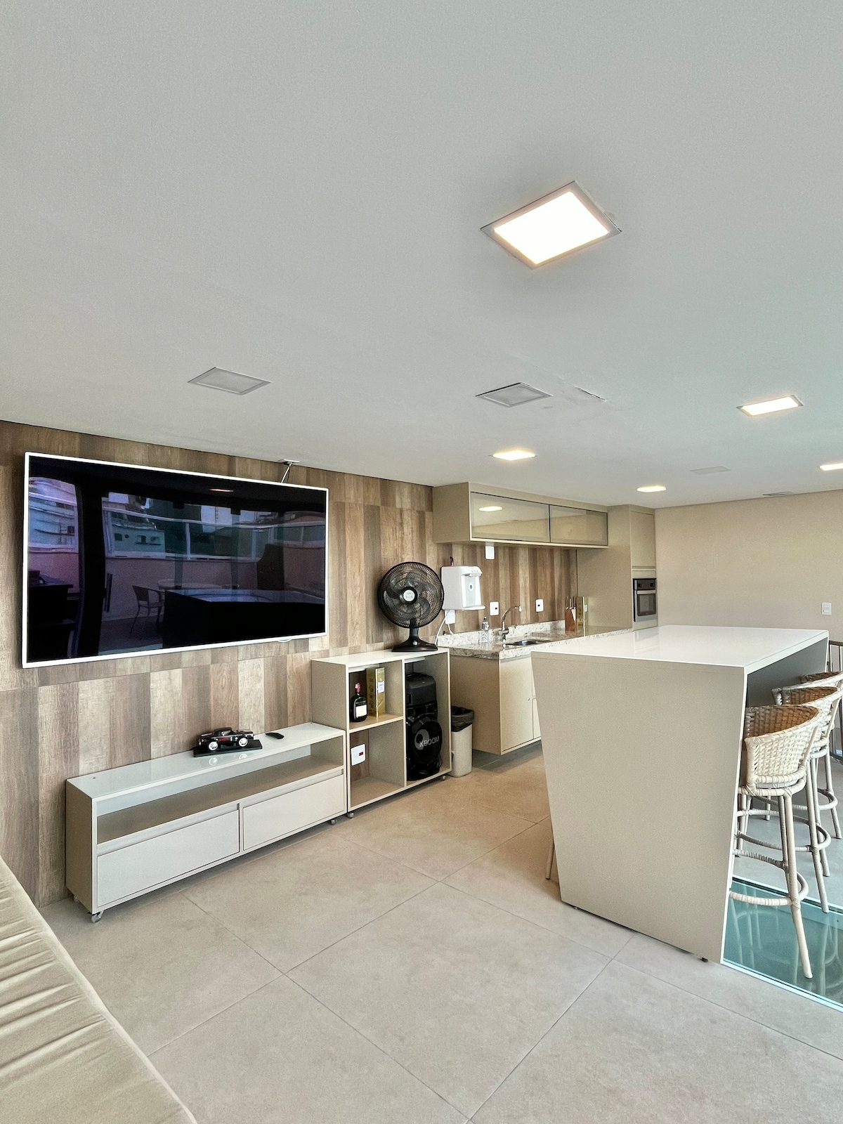 A modern living space is showcased, featuring a large wall-mounted television and a sleek kitchen area. Light-colored cabinets are paired with a white countertop, complemented by barstool seating. Natural light fills the room through large windows, enhancing the spacious atmosphere.