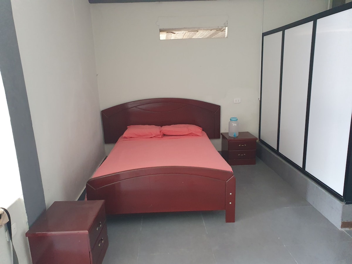 A spacious bedroom features a wooden bed with a coral-colored bedspread, accompanied by two matching nightstands. A simple glass container sits on one nightstand. Natural light filters in through a small window, and a partial room divider is visible to the right.