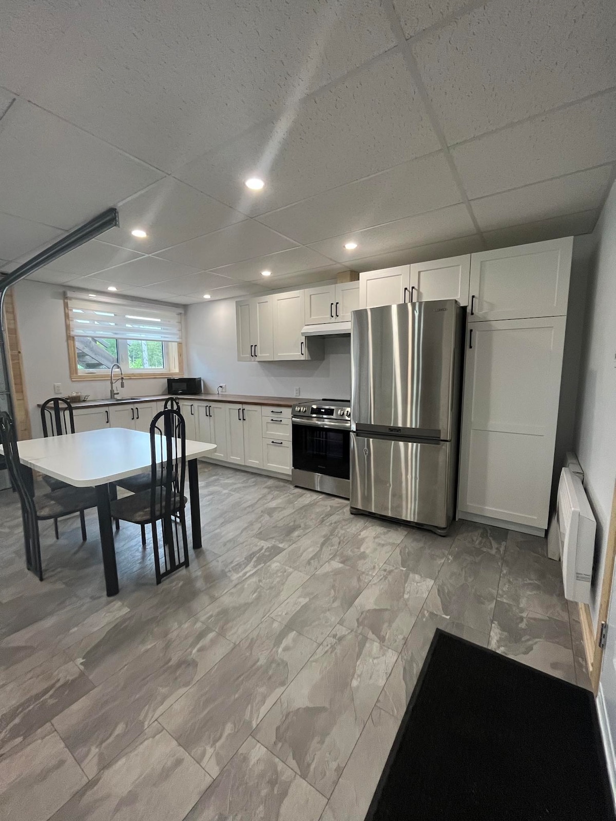 A modern kitchen is displayed with sleek white cabinetry and stainless-steel appliances. A dining table with four black chairs is positioned near a window, offering natural light. The floor features a light grey tile, enhancing the clean and spacious environment.