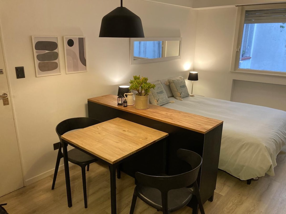 An open living space is presented, featuring a light-colored bed with multiple pillows. A wooden table with two black chairs is set in front of the bed, complemented by a small potted plant and a lantern. Two framed prints are displayed on the wall.