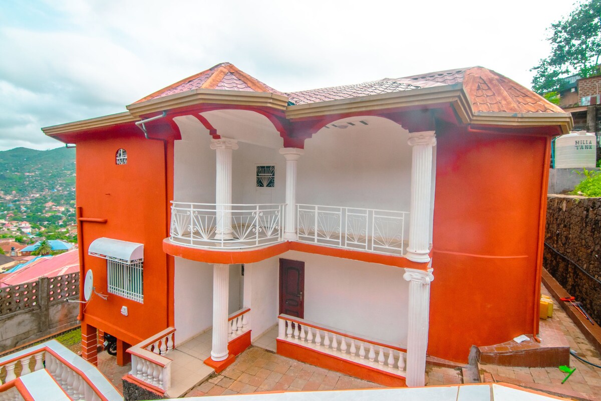 A vibrant orange exterior of a two-story guest house is highlighted, featuring a spacious terrace with white railings. The entrance is marked by an arched doorway, complemented by tall white columns. Lush greenery surrounds the property, nestled within a hillside neighborhood.