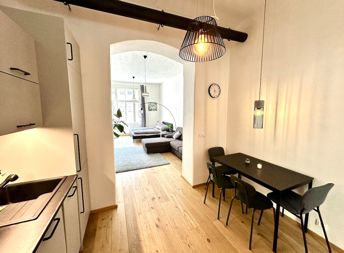 The open layout of the apartment is illustrated, featuring a modern kitchen that leads into a spacious living area. A black dining table with chairs is positioned near the kitchen, and a large sofa is visible in the living space, providing a welcoming environment.