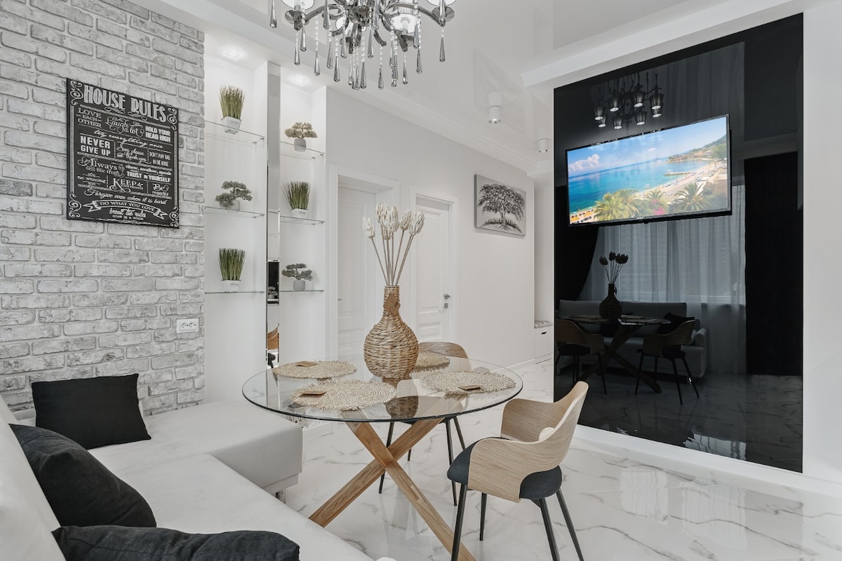 A modern living space is presented with a round glass table atop a light wooden base, surrounded by neutral-toned chairs. A large screen displays scenic views, while white walls are complemented by decorative elements and a stylish chandelier above.