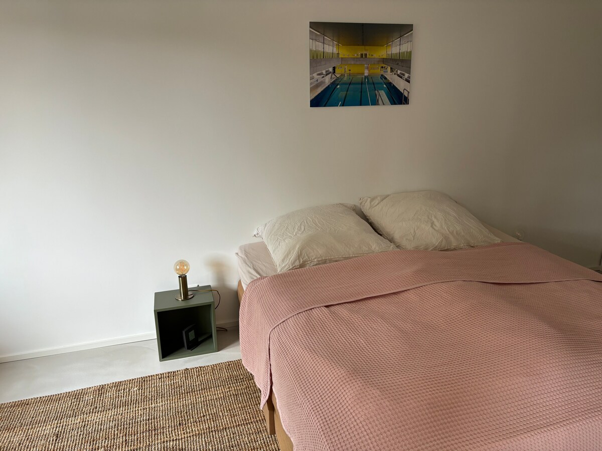 A spacious bed with soft pillows is covered by a light pink blanket. A compact nightstand holds a sleek lamp on one side. A serene artwork of a swimming pool hangs on the wall above the bed, contributing to the calming atmosphere of the room.