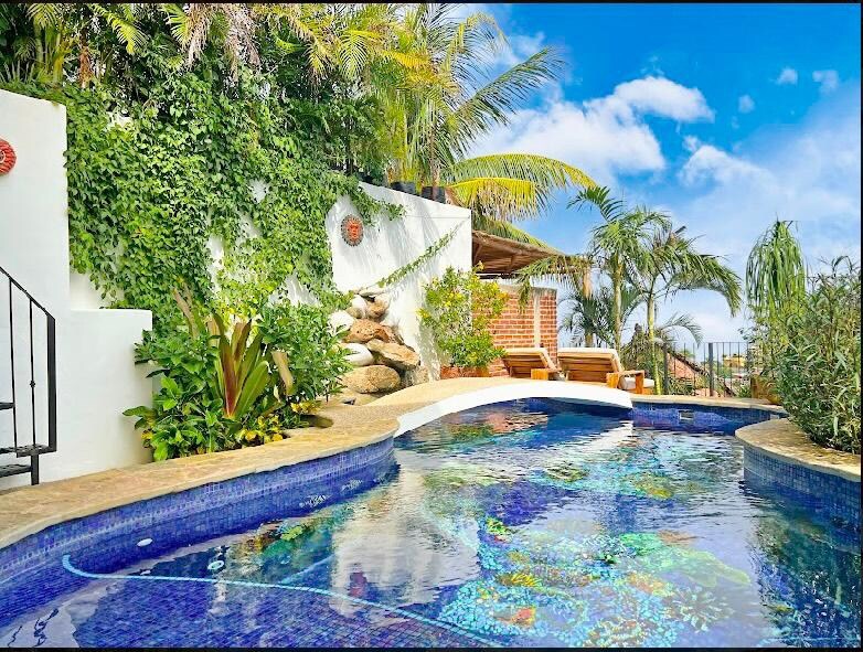 A private heated saltwater pool is surrounded by tropical greenery and palm trees, enhancing the serene atmosphere. Lounge chairs are positioned along the pool's edge, inviting relaxation. The bright blue sky adds to the inviting setting, complemented by stone and plant accents.
