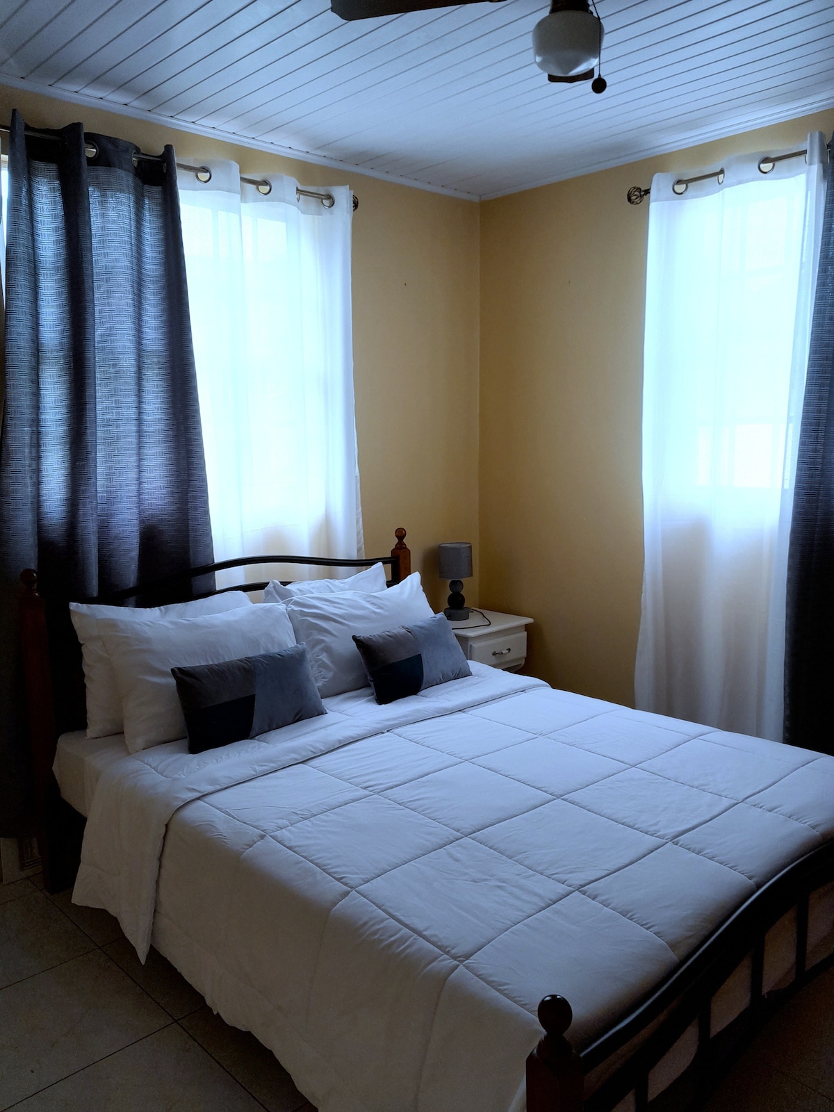 A cozy bedroom features a spacious bed dressed in crisp white linens with accent pillows. Light enters through two large windows, surrounded by curtains in contrasting colors. A bedside table and lamp are positioned next to the bed, contributing to a relaxing atmosphere.