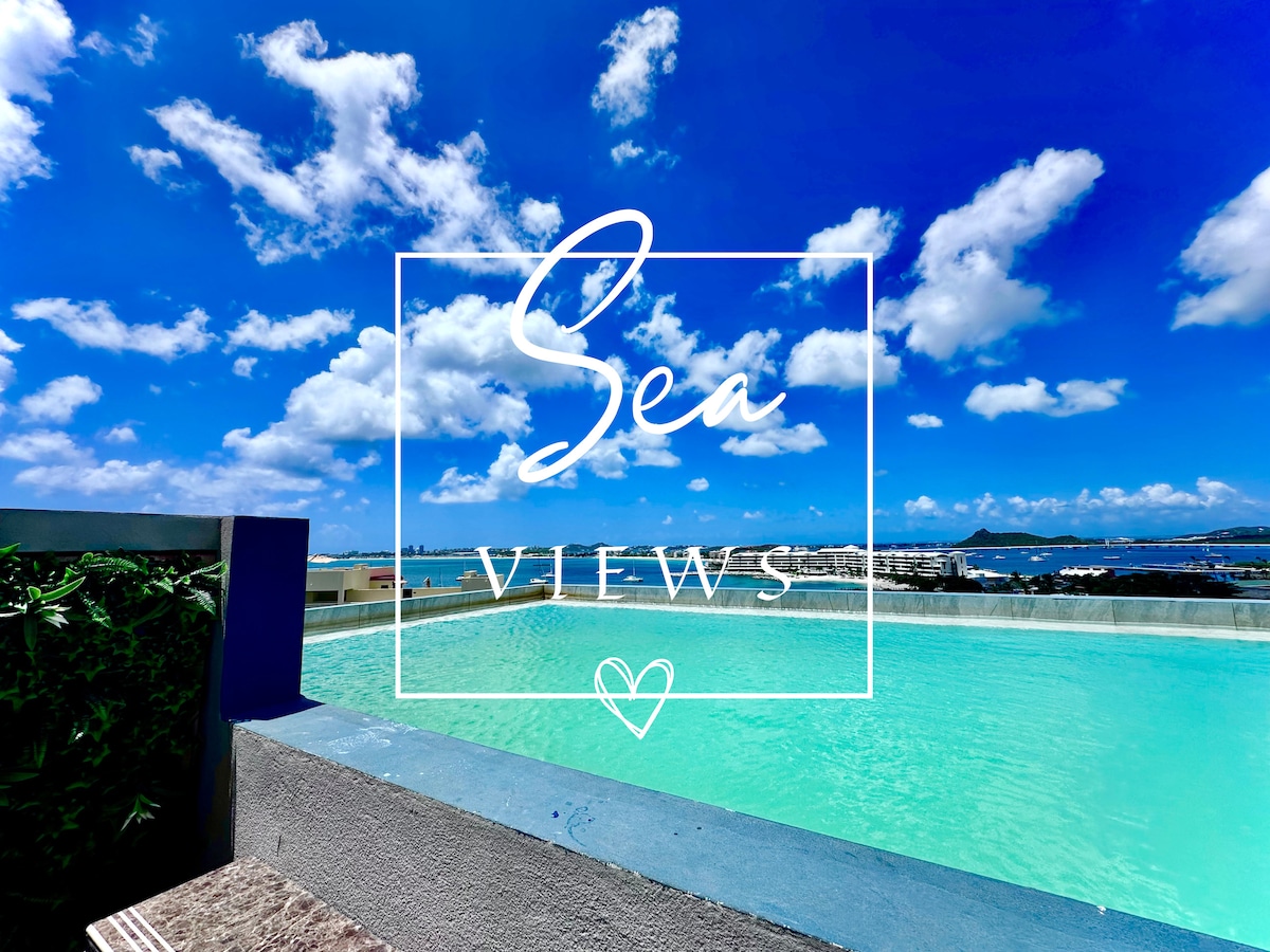 An expansive rooftop pool offers stunning views of the sea under a bright blue sky, accented by scattered clouds. The clear water reflects the vibrant scenery, providing a serene environment for relaxation and enjoyment.