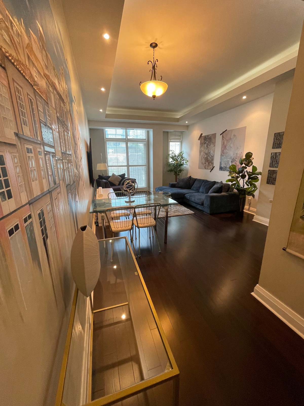 A spacious living area is highlighted by dark wood floors and abundant natural light. A large, comfortable couch is positioned next to a dining table with four chairs. A decorative console table is visible, enhancing the room's aesthetic with artwork adorning the walls.