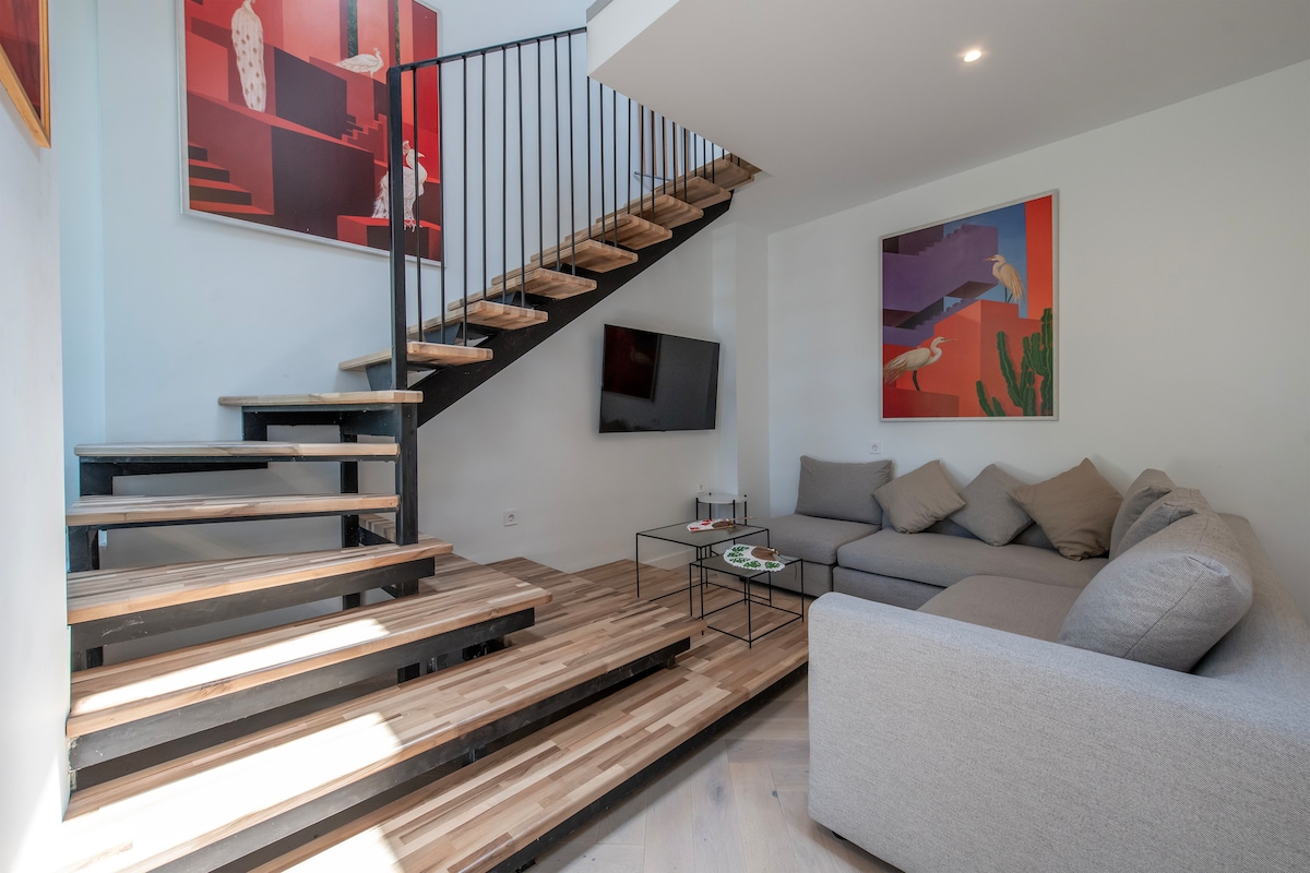 A modern living area is highlighted by a comfortable sectional sofa with neutral cushions. A minimalist coffee table sits nearby. A striking staircase with wooden steps and a metal railing leads to the upper level. Colorful artwork adorns the walls, adding interest to the space.