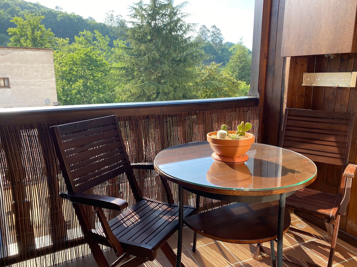 A bright terrace area features a round glass table accompanied by two wooden chairs. A potted plant with vibrant greenery occupies the center of the table. Lush trees and hills provide a scenic backdrop, enhancing the outdoor space.
