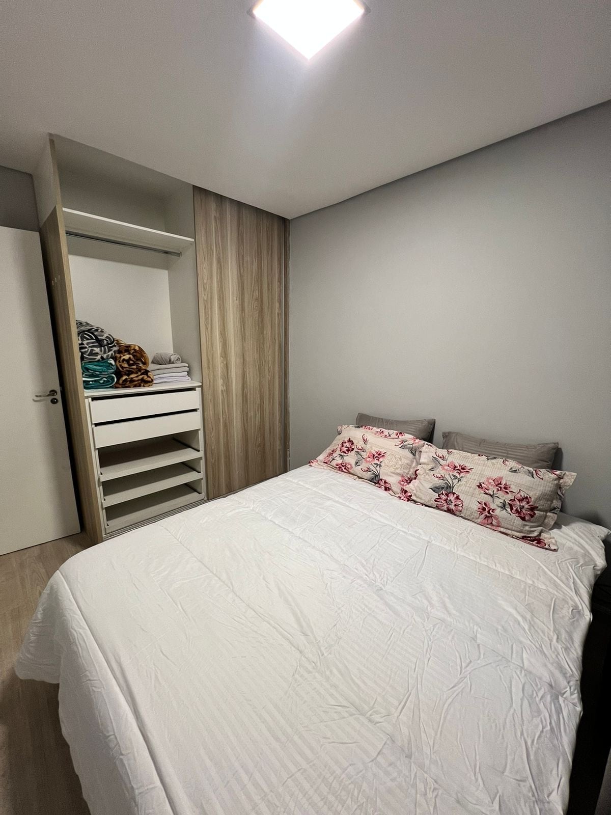 A sleeping area features a spacious bed with a light-colored duvet and floral-patterned pillows. A closet with neatly organized shelves and folded towels is visible, complementing the soft gray walls and warm wooden accents.