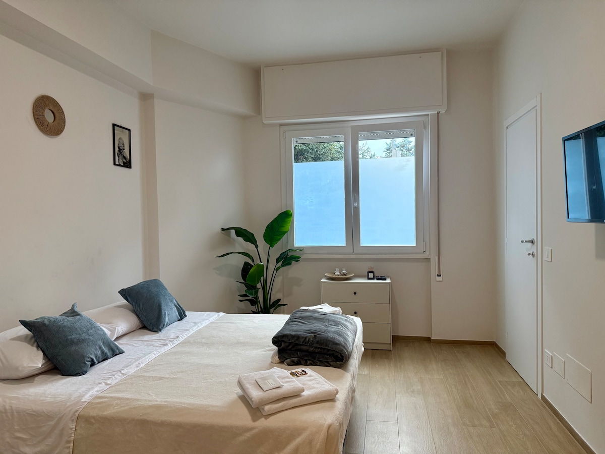A spacious bedroom features a king-size bed dressed in white linens, accompanied by two decorative pillows. A tall indoor plant stands beside the bed. Natural light enters through large windows, and a minimalist dresser is located across from the bed, with a wall-mounted TV nearby.