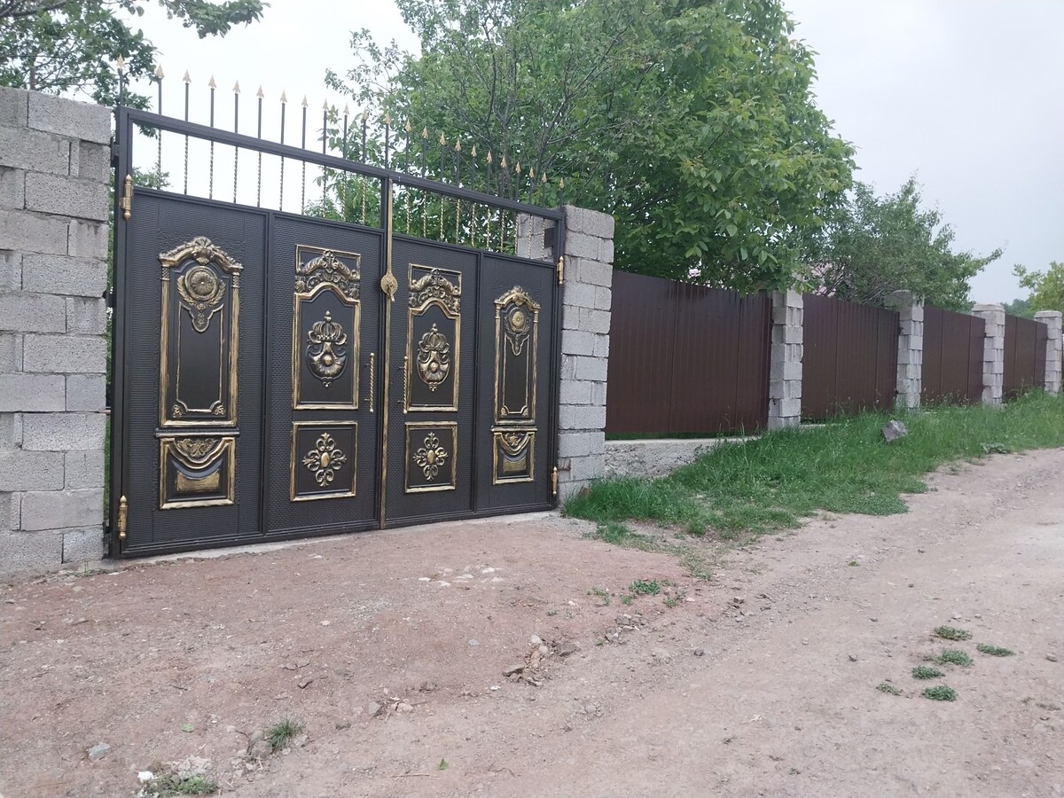 An ornate black and gold gate is set against a backdrop of a low stone wall and a wooden fence. Lush greenery, including trees and grass, is visible nearby, enhancing the entrance to the property.