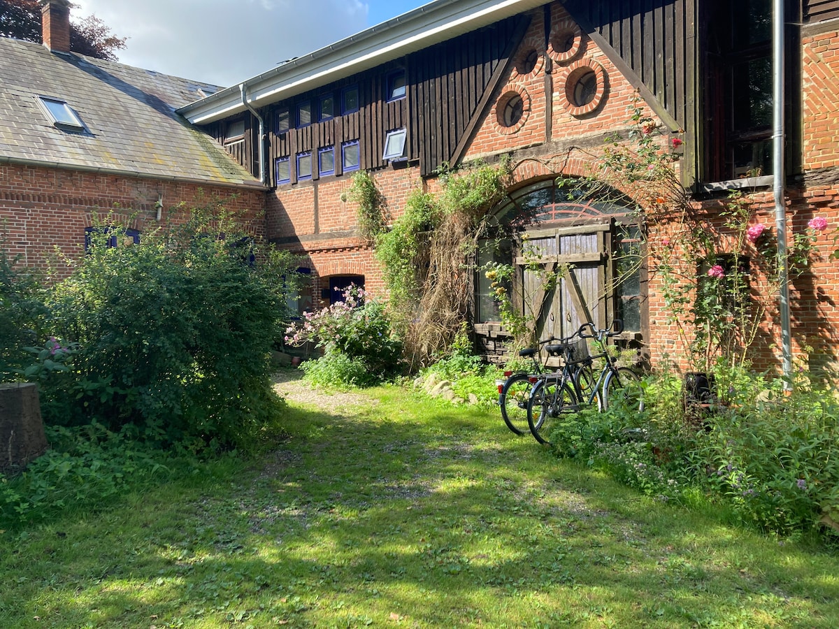 A charming outdoor space is featured, surrounded by lush greenery and blooming flowers. A pathway leads toward a historic wooden door, framed by ivy and a rustic brick facade. Two bicycles are parked on the grass, adding to the inviting atmosphere of the garden.