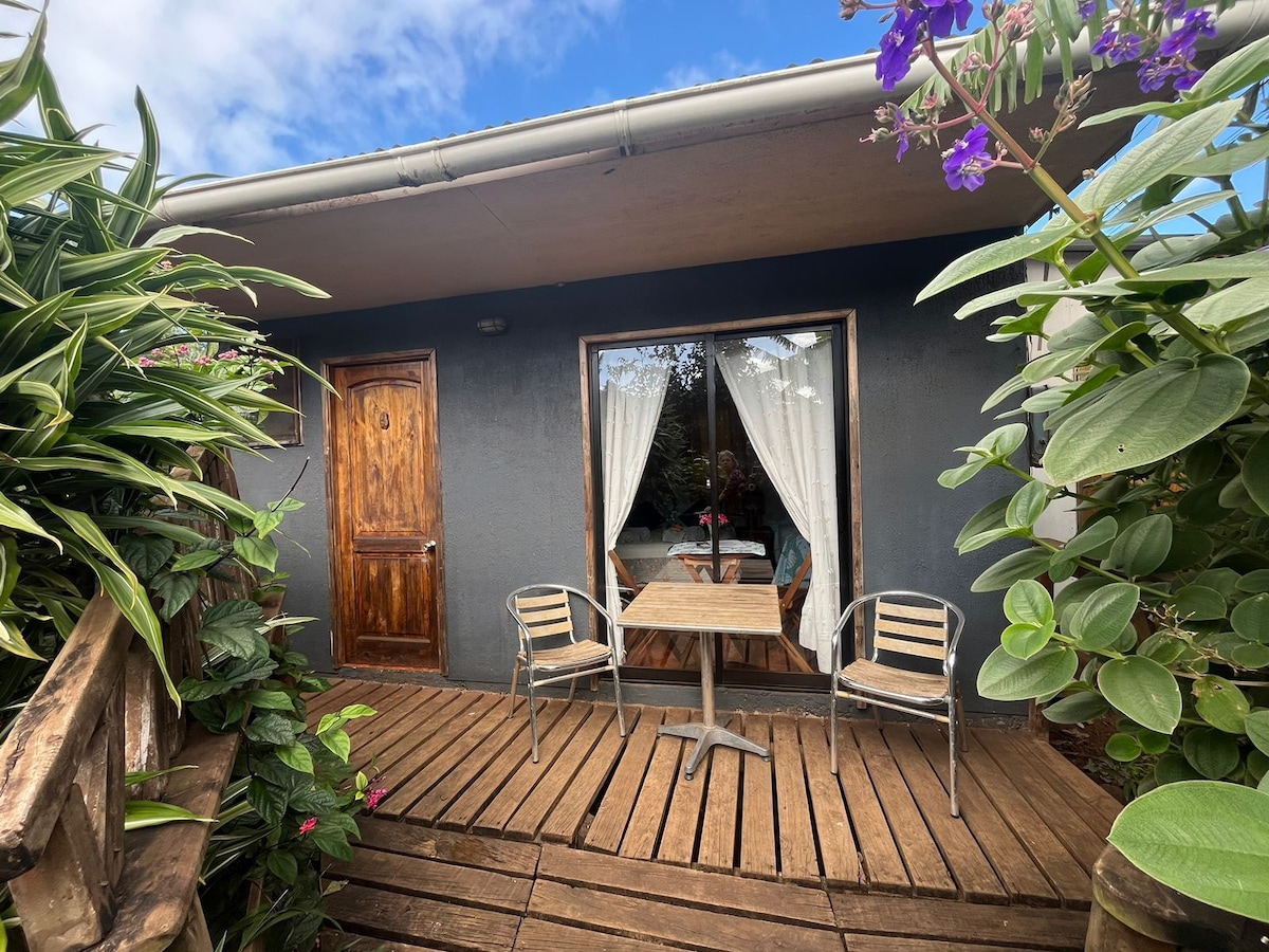 A private terrace features a wooden deck and two metal chairs surrounding a small table. Lush greenery and colorful flowers frame the entrance, while tall curtains softly filter light from within the cabin, enhancing the serene atmosphere of the outdoor space.