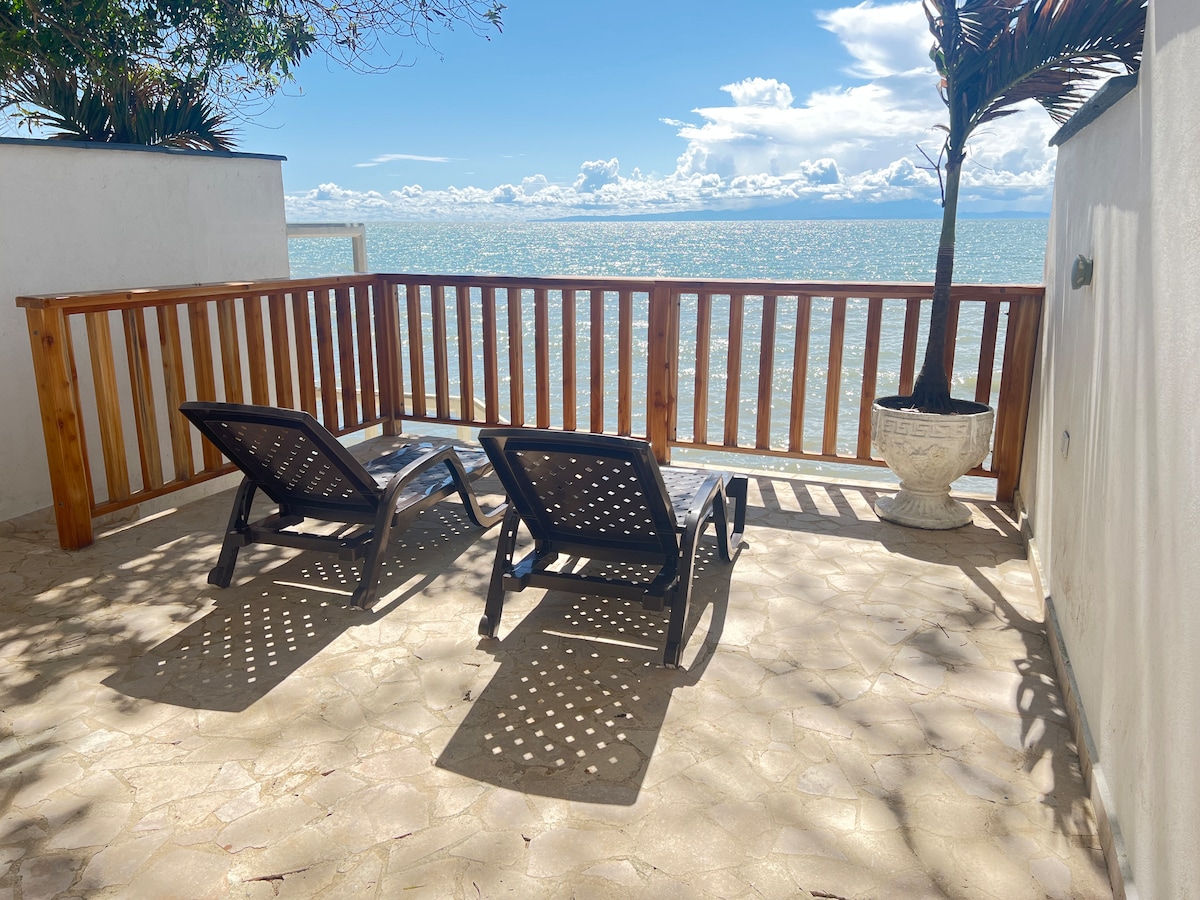 Two lounge chairs are positioned on a private terrace overlooking the sea, framed by a wooden railing. A large plant adds greenery to the space, with sunlight casting playful shadows on the textured stone floor. The horizon features sparkling water under a clear sky.